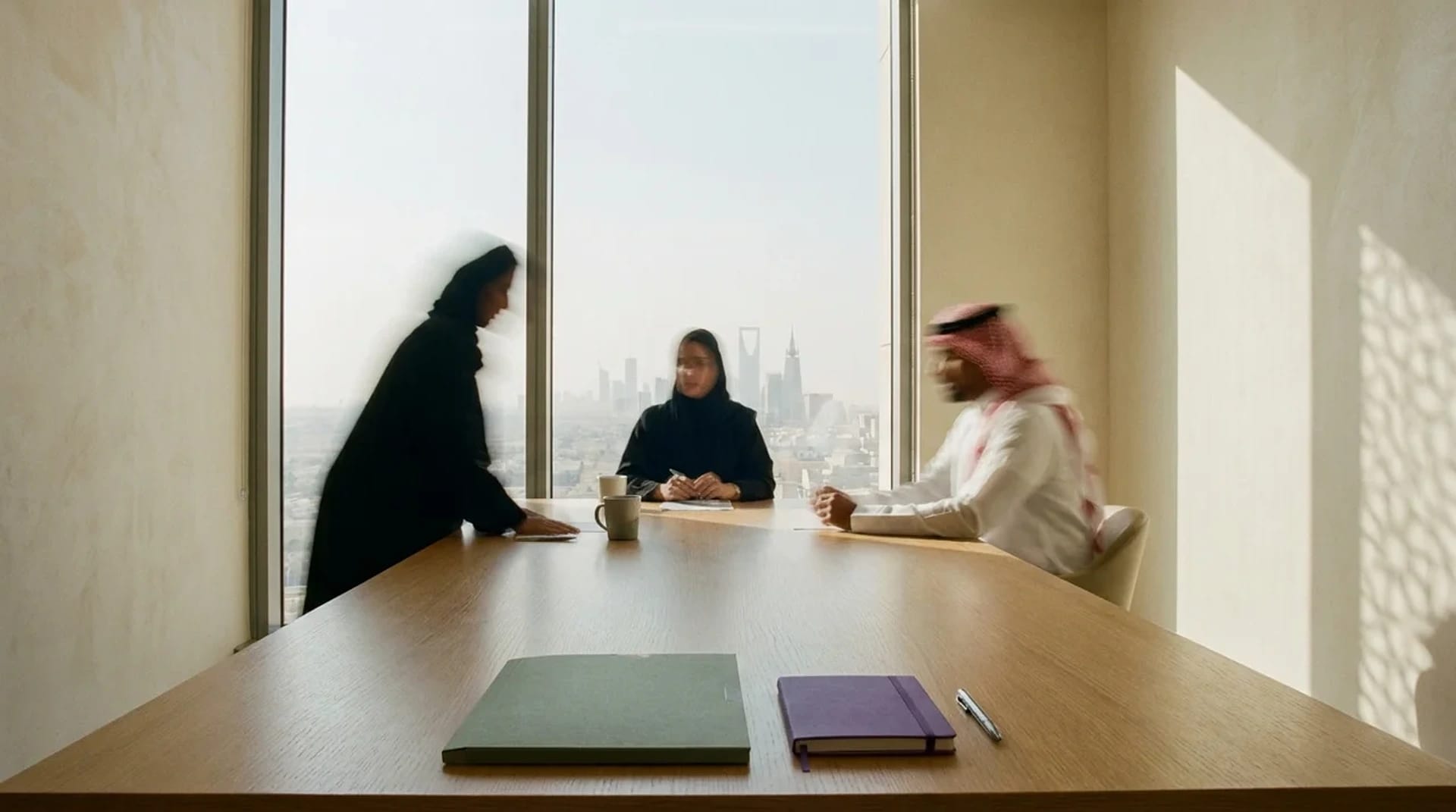 A consulting session around a meeting table overlooking the Riyadh skyline — the room still, the people in motion